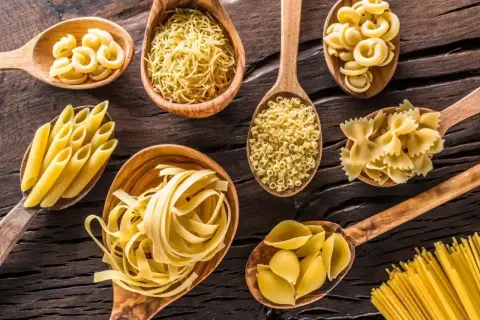 Drewniane łyżki pełne różnorodnego makaronu: penne, farfalle, muszelki, spaghetti i inne. Każdy rodzaj to inna porcja makaronu ugotowanego kcal.