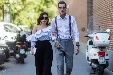 Pareja elegante pasea, él es un invitado boda hombre sin traje, con tirantes y tatuajes. Ella lleva un top blanco voluminoso.