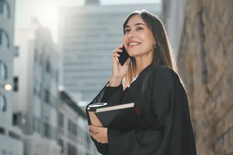 Uśmiechnięta prawniczka w todze rozmawia przez telefon, trzymając książkę i kawę.