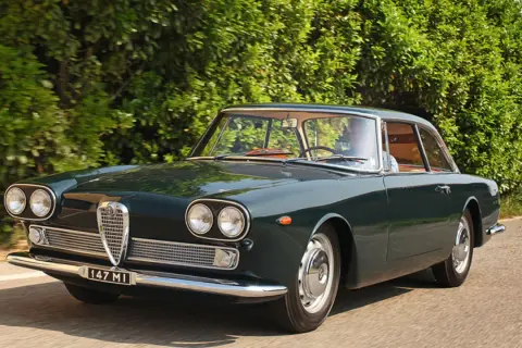En mörkgrön Alfa Romeo Giulia Sprint GT kör längs en landsväg, en klassisk skönhet bland Alfa Romeo modeller.