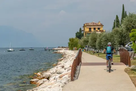 Ciclista sul lungolago, con barche e montagne sullo sfondo. Un assaggio di cosa vedere sul Monte Baldo, tra natura e relax.