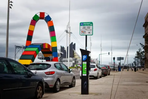 Panneaux de stationnement payant et horodateur, rappelant qu'une amende stationnement non payé est possible. Au loin, une arche colorée.