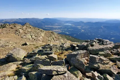 Vista panorámica de los picos de la Sierra de Guadarrama, con rocas cubiertas de líquenes en primer plano y un valle con un lago al fondo.