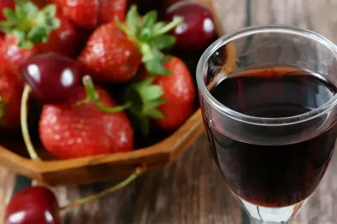 Szklanka ciemnego płynu, prawdopodobnie ratafia wódka, obok miski pełnej świeżych truskawek i wiśni.