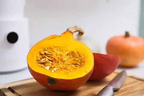 Pomarańczowa dynia przekrojona na pół, pełna nasion, obok inne odmiany dyni i nóż na desce.
