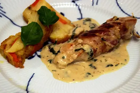 En kycklingrulle insvept i bacon, serverad med en krämig sås till kyckling och gratinerade potatisbitar med gröna blad.