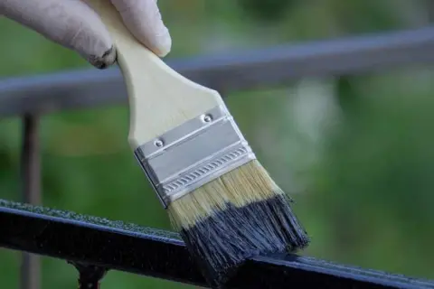 Mano guantata che inizia a verniciare ringhiera con pennello nero.