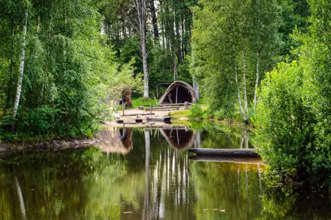 En stilla sjö speglar grönskande träd och en hydda vid Årås kvarn. En stockbåt vilar vid strandkanten.
