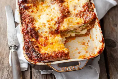 Lasagne alla bolognese appena sfornate, dorate e filanti, in una teglia Staub. Un coltello vintage è appoggiato accanto.