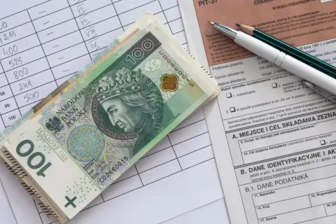 Stos banknotów 100 zł leży na formularzu PIT. Obok arkusz z danymi i ołówki.
