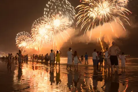 Ludzie na plaży podziwiają spektakularny pokaz fajerwerków. To idealny sylwester w ciepłych krajach, z odbiciami świateł w wodzie.