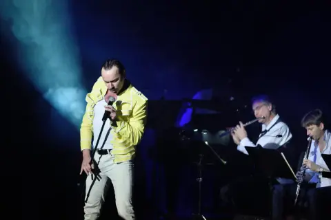 Wokalista w żółtej kurtce śpiewa na scenie, podczas gdy muzycy grają na instrumentach dętych. To koncert queen symfonicznie.