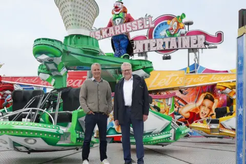 Zwei Männer stehen vor einer bunten Kirmesattraktion mit Clowns und der Aufschrift "Entertainer". Ein fröhlicher Ausblick auf das Bayreuth Herbstfest.
