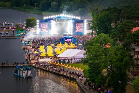Tłumy ludzi na festiwalu Rap Stacja, scena z ekranami i dymem, żółte parasole, łódź na wodzie.