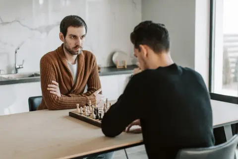 Dwóch mężczyzn gra w szachy. Jeden z nich, w brązowym swetrze, zamyślony patrzy na planszę, jakby zastanawiał się nad ruchem króla.