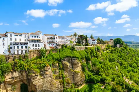 Vita hus i Ronda, Spanien, klättrar uppför en grönskande klippa under en blå himmel med moln.
