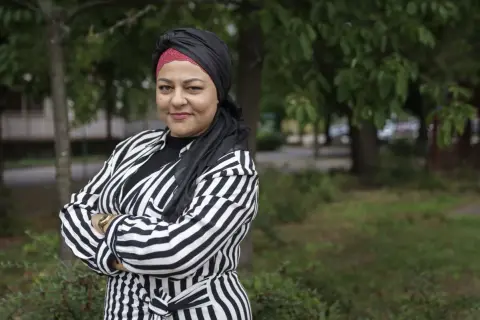Zeina Mourtada, med en svart och röd turban och en randig jacka, står med armarna i kors utomhus.