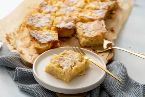 Ein saftiger Apfelkuchen vom Blech, bestreut mit Puderzucker und Mandelblättchen. Ein Stück liegt auf einem Teller, bereit zum Genuss.