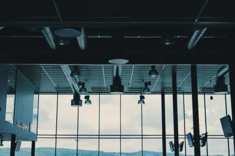 Synliga ventilationsrör och belysning i taket, med stora fönster som visar en molnig himmel och en grönskande kulle.