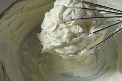 Un fouet de cuisine retient une généreuse portion de glacage mascarpone crémeux, prêt à garnir un gâteau.