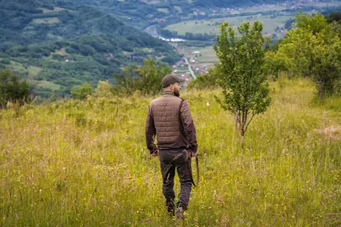 Uomo con fucile attraversa prati fioriti, godendosi la vista dei passi di montagna e della valle sottostante.