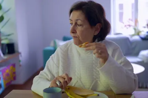 Kobieta w białym swetrze je banana, czując uczucie guli w gardle. Na stole kubek i talerz.