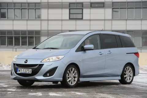 Jasnoniebieska Mazda 5 z nowymi żarówkami w reflektorach, zaparkowana przed nowoczesnym budynkiem.
