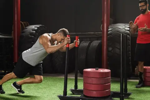 Mężczyzna wykonuje ćwiczenie sled push, pchając obciążony sprzęt. Trener obserwuje jego wysiłek.