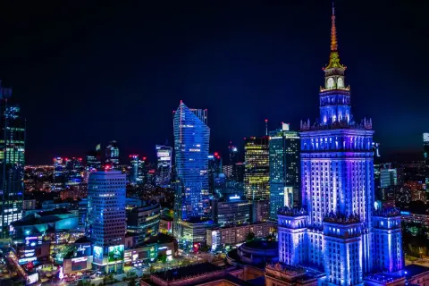 Nocna panorama Warszawy z Pałacem Kultury oświetlonym na niebiesko. Miasto tętni życiem, a liczne światła zapowiadają ciekawe warszawa wydarzenia.