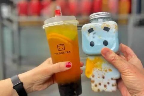 Zwei Hände halten leckere Getränke: ein Bubble Tea von Mr. Box Tea in Berlin und ein süßer blauer Drink in einer Figurenflasche.