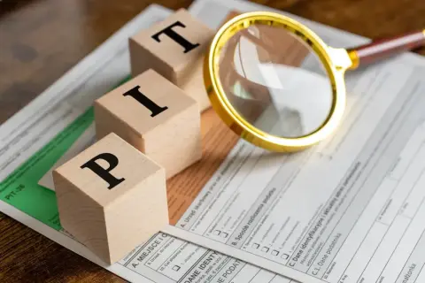 Drewniane klocki z literami "PIT" leżą na formularzu podatkowym. Złota lupa przybliża szczegóły. Czas zacząć myśleć, od kiedy można rozliczać PIT.