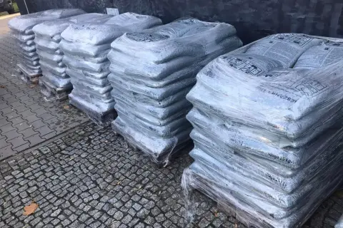 Stosy worków z asfaltem na paletach, owinięte folią, na kamiennym podłożu.