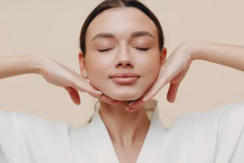 Mujer con ojos cerrados, disfrutando de un masaje facial. Sus manos sostienen suavemente su rostro, practicando yoga facial para una piel radiante.