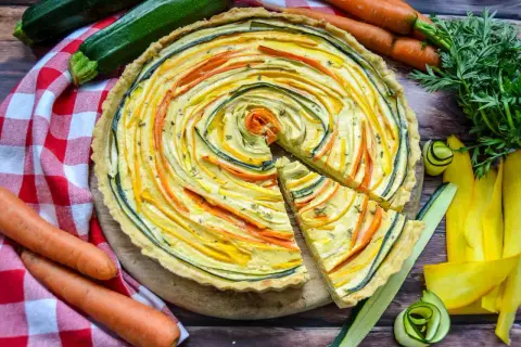 Bunte quiche vegetarisch mit Zucchini und Karotten, kunstvoll im Kreis arrangiert. Daneben frisches Gemüse.