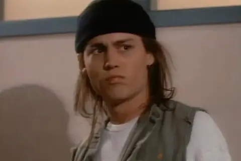 Johnny Depp ung med långt hår och svart mössa, bär en väst över en vit tröja.