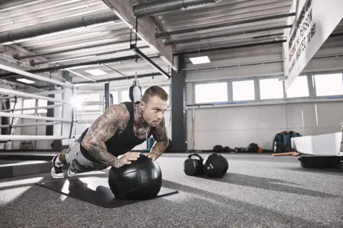 Mann mit Tattoos macht Liegestütze auf einem Medizinball, fokussiert auf sein hypertrophie training. Im Hintergrund ein Boxring und Trainingsgeräte.