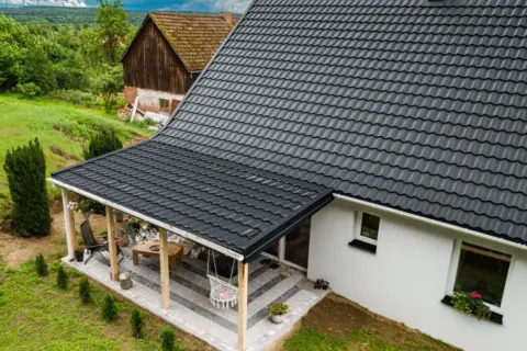 Nowy, ciemnoszary dach z blachodachówki nad tarasem i domem, otoczony zielenią.