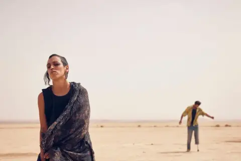 Mujer con pañuelo en desierto, hombre con muleta al fondo. El sirat, significado de un camino difícil.