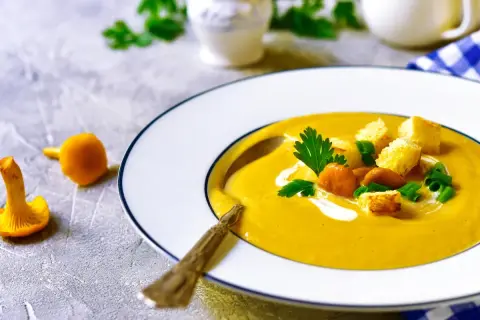 Aksamitna zupa krem z kurek, udekorowana grzankami, świeżą natką pietruszki i szczypiorkiem. Obok talerza leżą świeże kurki.