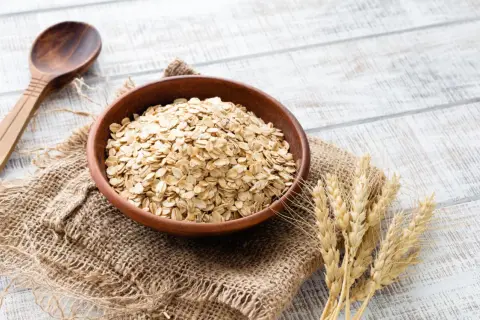 Schüssel mit Haferflocken, die für ihren guten Haferflocken proteingehalt bekannt sind, daneben eine Holzschale und Weizenähren.