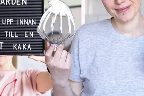 En person håller upp en skylt med texten "Ge inte upp innan du fått till en perfekt kaka". Bredvid syns en visp med grädde och en tom burk, redo för en vegansk fika.