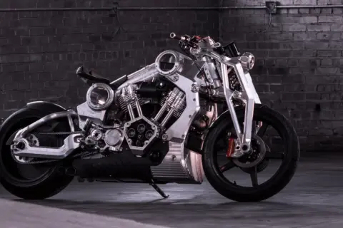 Wyjątkowy, lśniący motocykl, prawdopodobnie najdroższy motor na świecie, z masywnym silnikiem i detalami z polerowanego metalu, na tle ceglanej ściany.