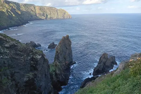 Acantilados escarpados y rocas emergiendo del mar azul en la **costa Artabra**. El cielo nublado ilumina el paisaje salvaje.