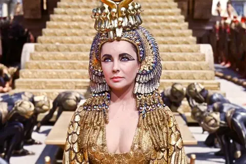 Elizabeth Taylor jako Kleopatra w złotej, bogato zdobionej sukni i koronie, z poddanymi klęczącymi u stóp.