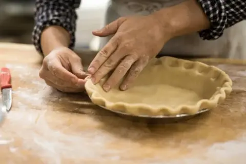 Mains farinées façonnant une pâte à tarte dans un moule. L'ajout d'une touche d'ammonium bicarbonate assure une croûte légère et croustillante.