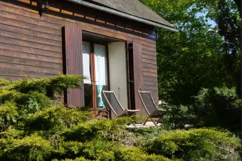 Chalet en bois avec étang de pêche privé en Franche-Comté. Deux chaises longues sur la terrasse, entourées de verdure luxuriante.