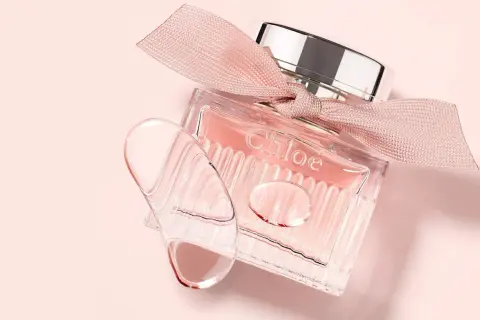 Szklana buteleczka L'eau de Chloe z różową wstążką i kroplami perfum na delikatnym, różowym tle.