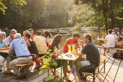 Menschen genießen das Leben im cafe am neuen see. Sie sitzen an Holztischen, trinken Bier und unterhalten sich. Im Hintergrund fließt ein ruhiger Fluss.