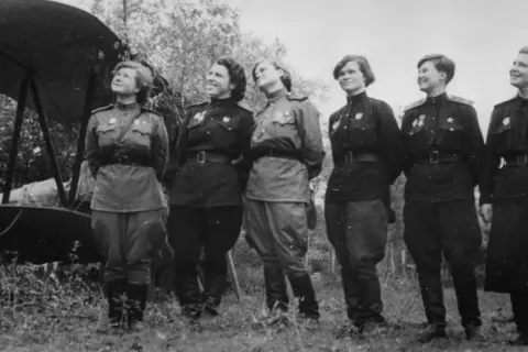 Seis mujeres con uniformes militares posan junto a un biplano. Son las brujas de la noche, listas para volar.