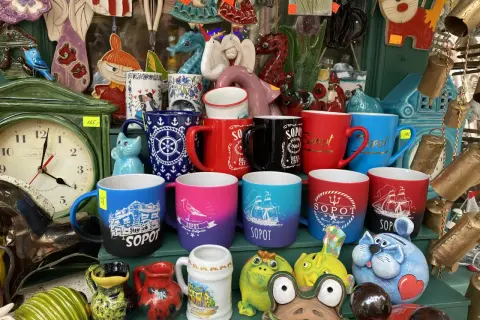 Kolekcja kubków z napisami "Sopot" i morskim motywem, idealne pamiątki z wakacji. W tle zegar i ceramiczne figurki.